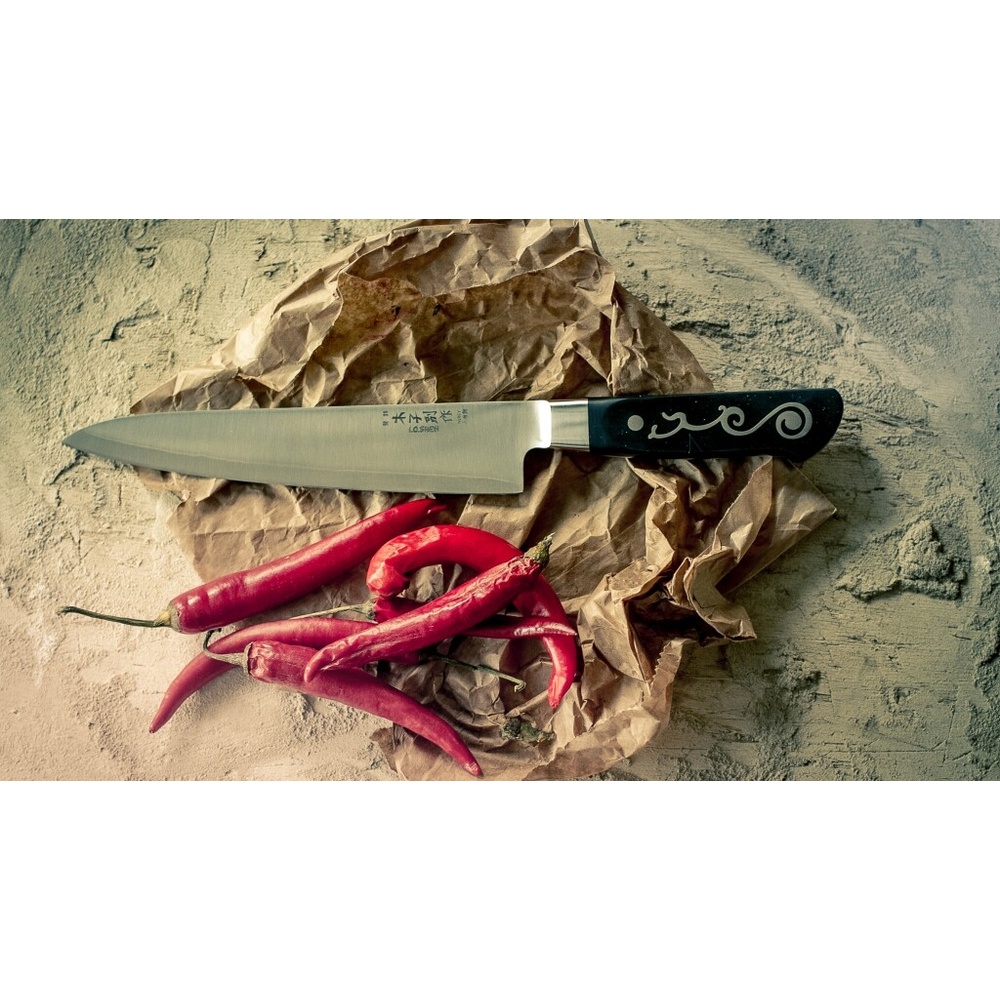 Large kitchen knife with black handle on crumpled brown paper, beside several red chili peppers on a textured surface.