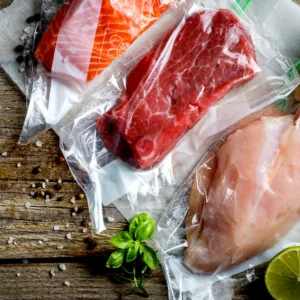 Vacuum-sealed raw beef and chicken on a wooden surface with herbs nearby (basil) and a lime wedge.