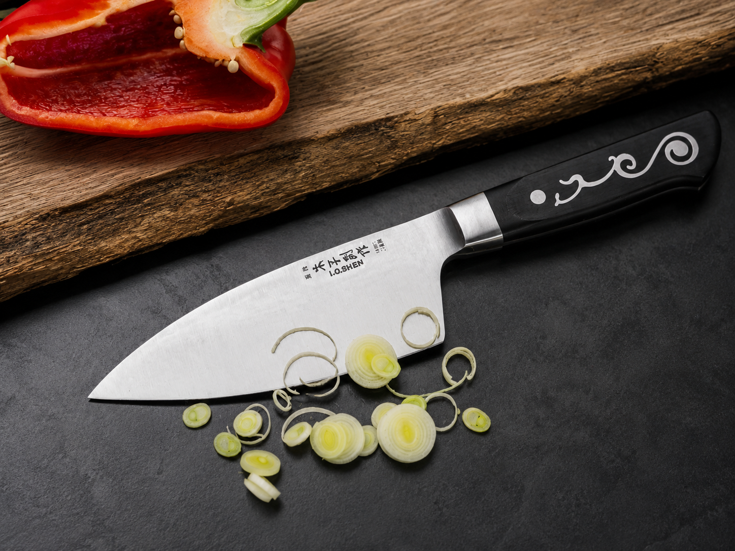 Chef's knife with black handle and decorative pattern rests on a dark surface, with sliced leeks and a red pepper on a wooden board.