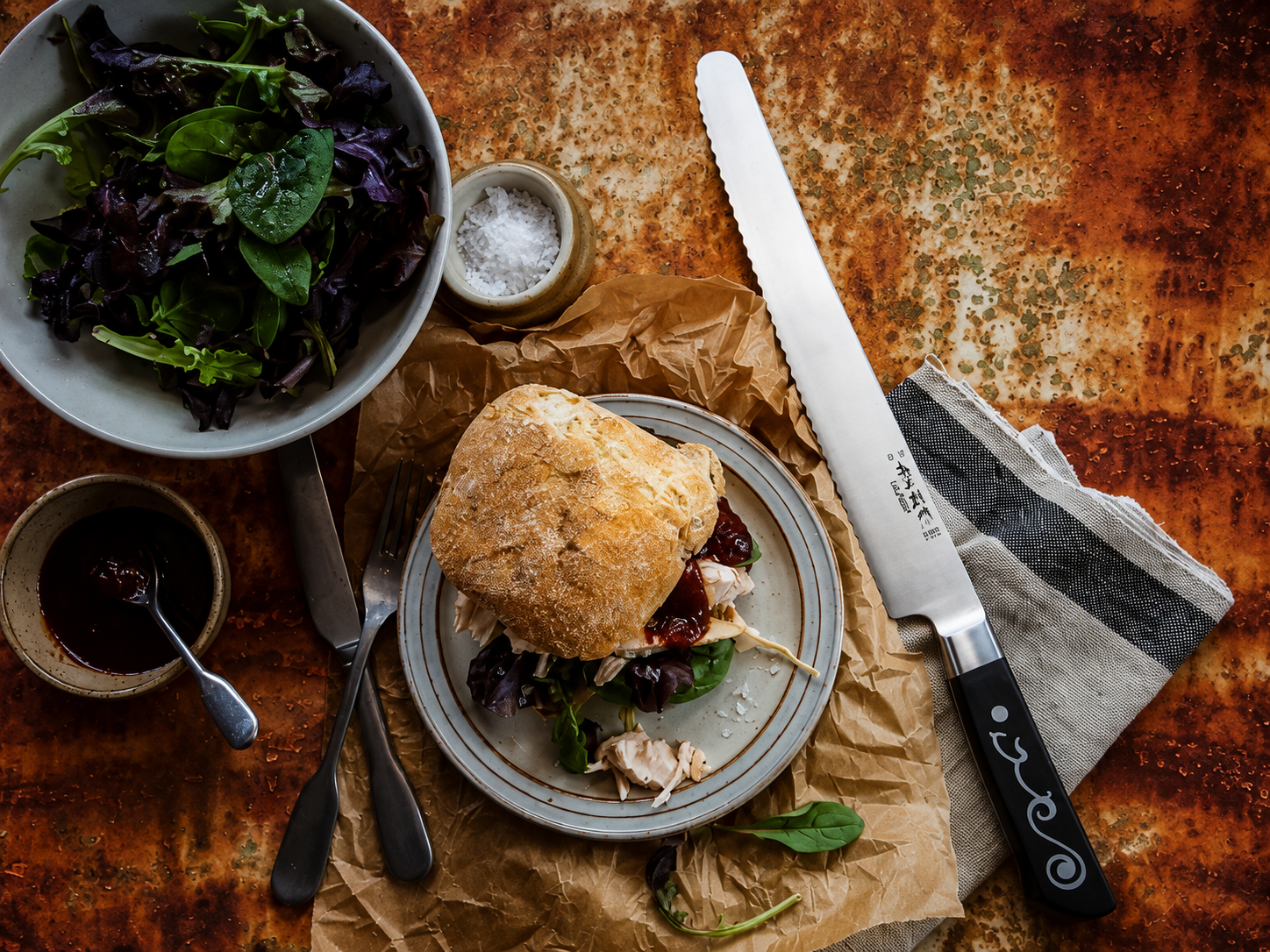 Rocky rustic table with a crusty sandwich on a plate, greens peeking from beneath, plus a side salad bowl and dipping sauce nearby