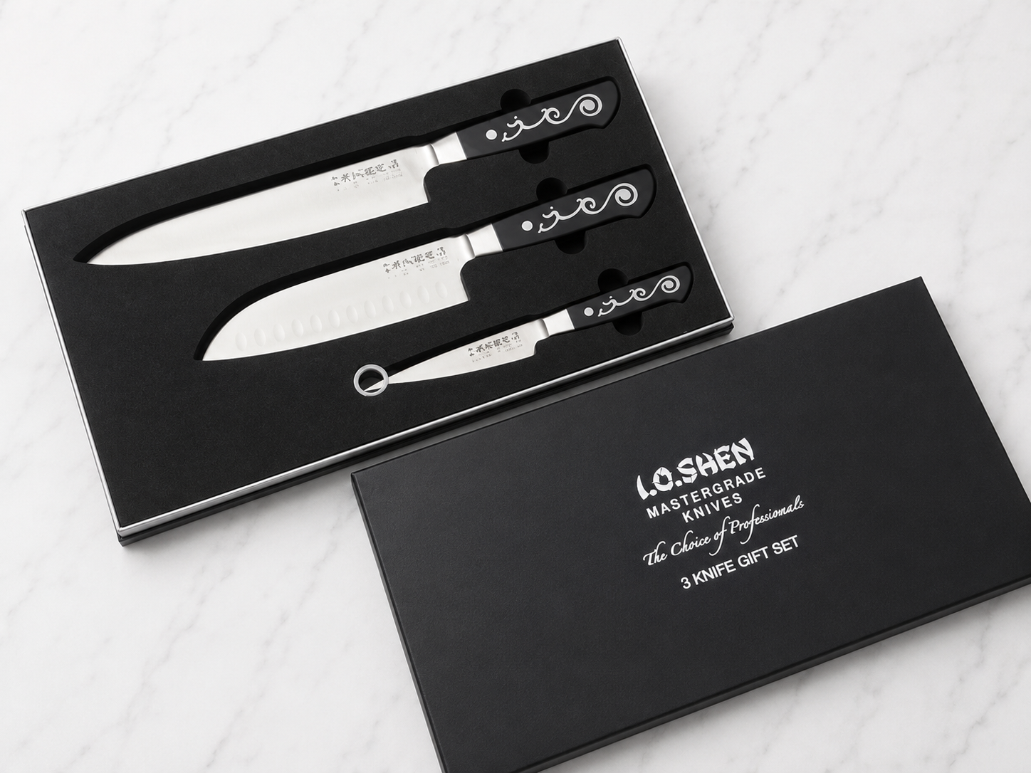 Three stainless steel kitchen knives with black decorative handles in a black foam insert inside a black gift box on a marble surface.