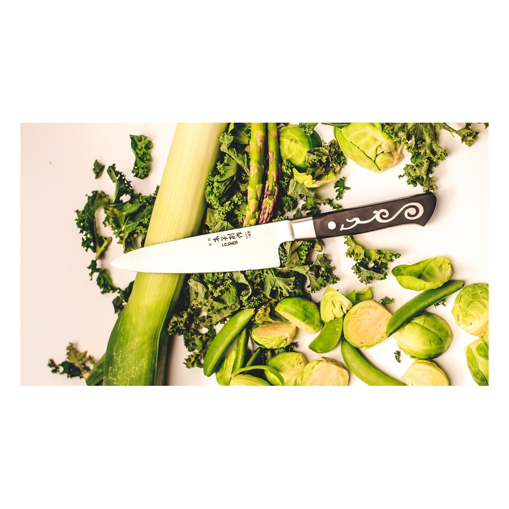 Chef's knife resting on a bed of kale with a leek, Brussels sprouts and snap peas scattered around on a white surface.