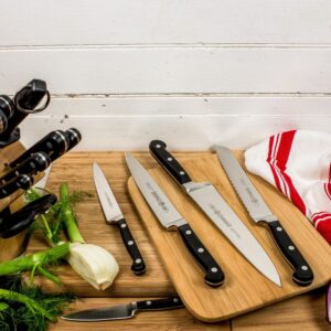 Kitchen knife set laid out on a wooden cutting board with fresh fennel and green onions nearby, and a red-and-white towel in the background.