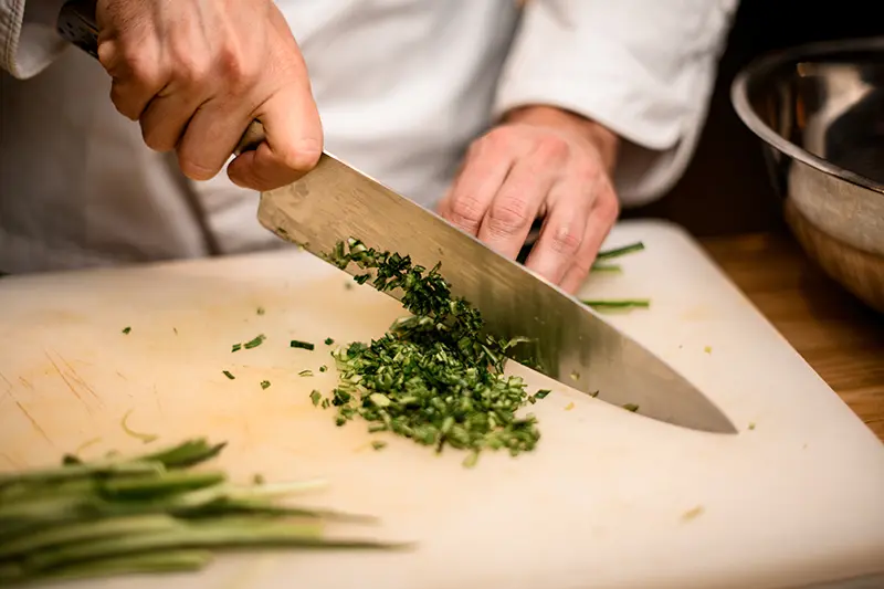 Chef’s knife sharpening - WA Hospitality Supply Hands mincing fresh green herbs with a chef's knife on a cutting board.